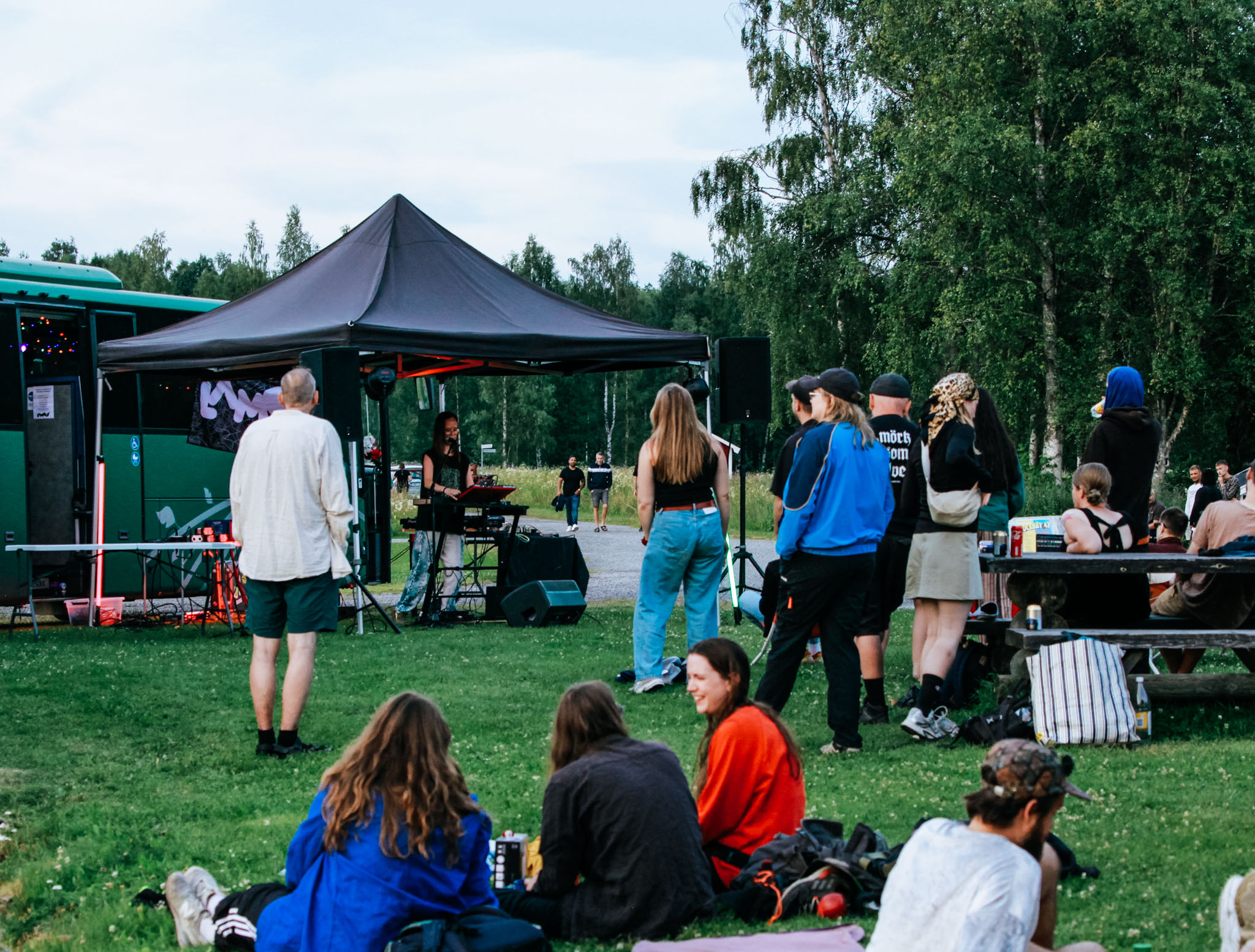 Festivalbesökare som sitter på en bänk och på marken. Under ett tälttak står en ung tjej och verkar DJa.
