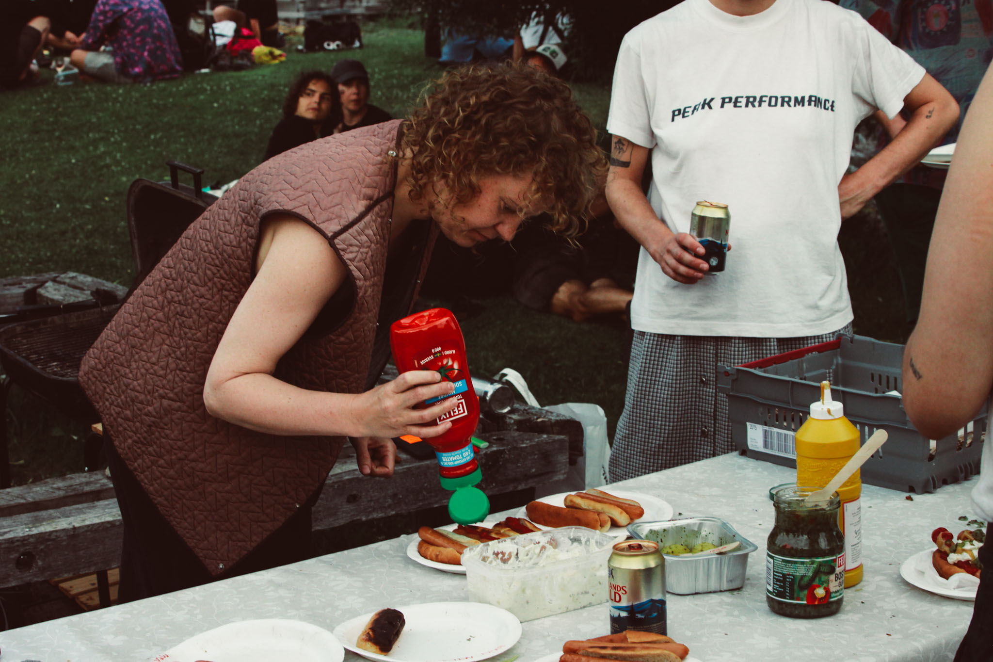 En kvinna häller ketchup på en tallrik med korv och bröd. På bordet finns även bostongurka, senap och två baljor med någon sås i. Bredvid kvinnan står någon i rutig kjol och vit t-shirt och håller en ölburk.