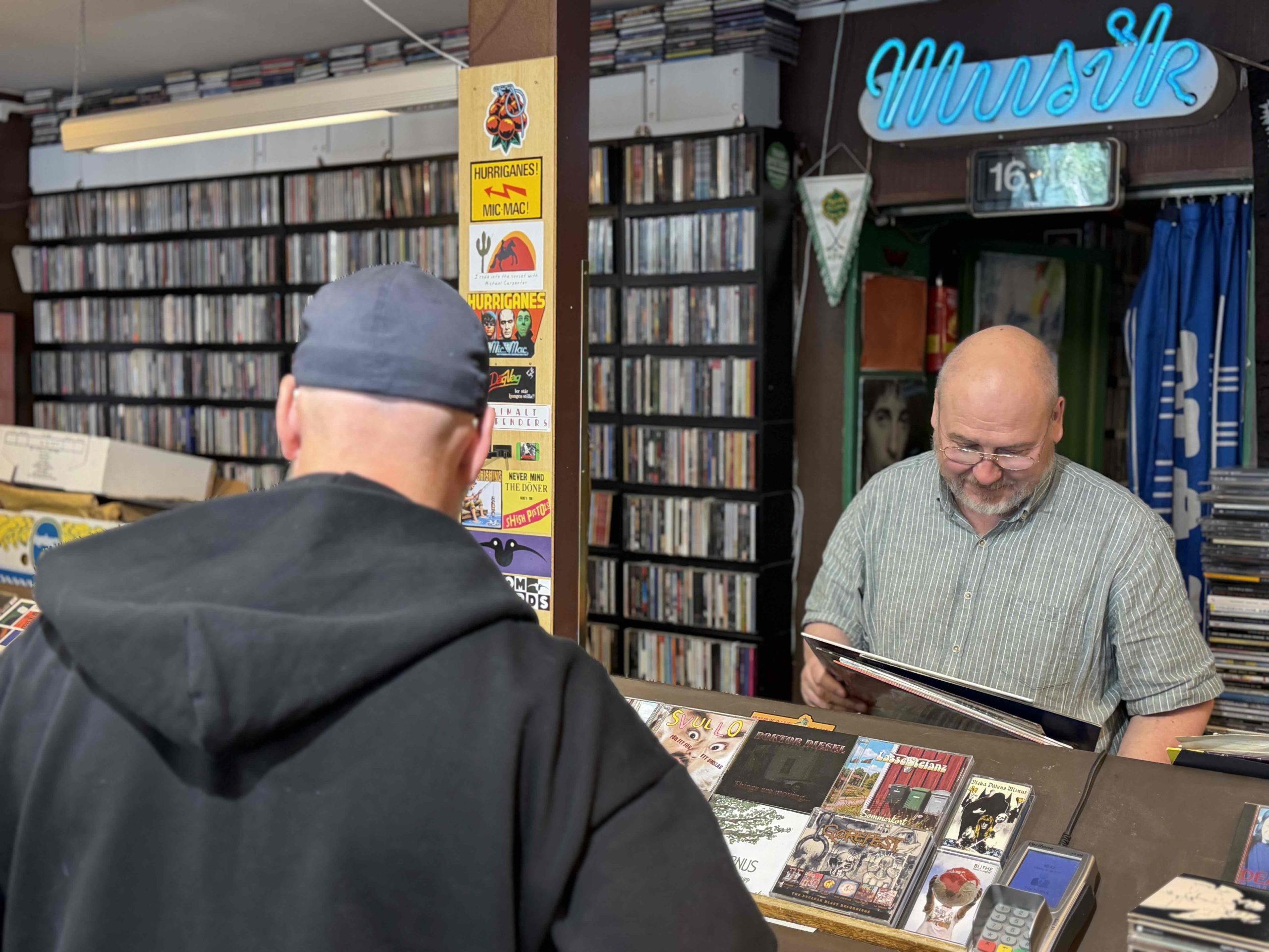 Butikens ägare läser på baksidan av en vinylskiva. Framför honom står en kund.