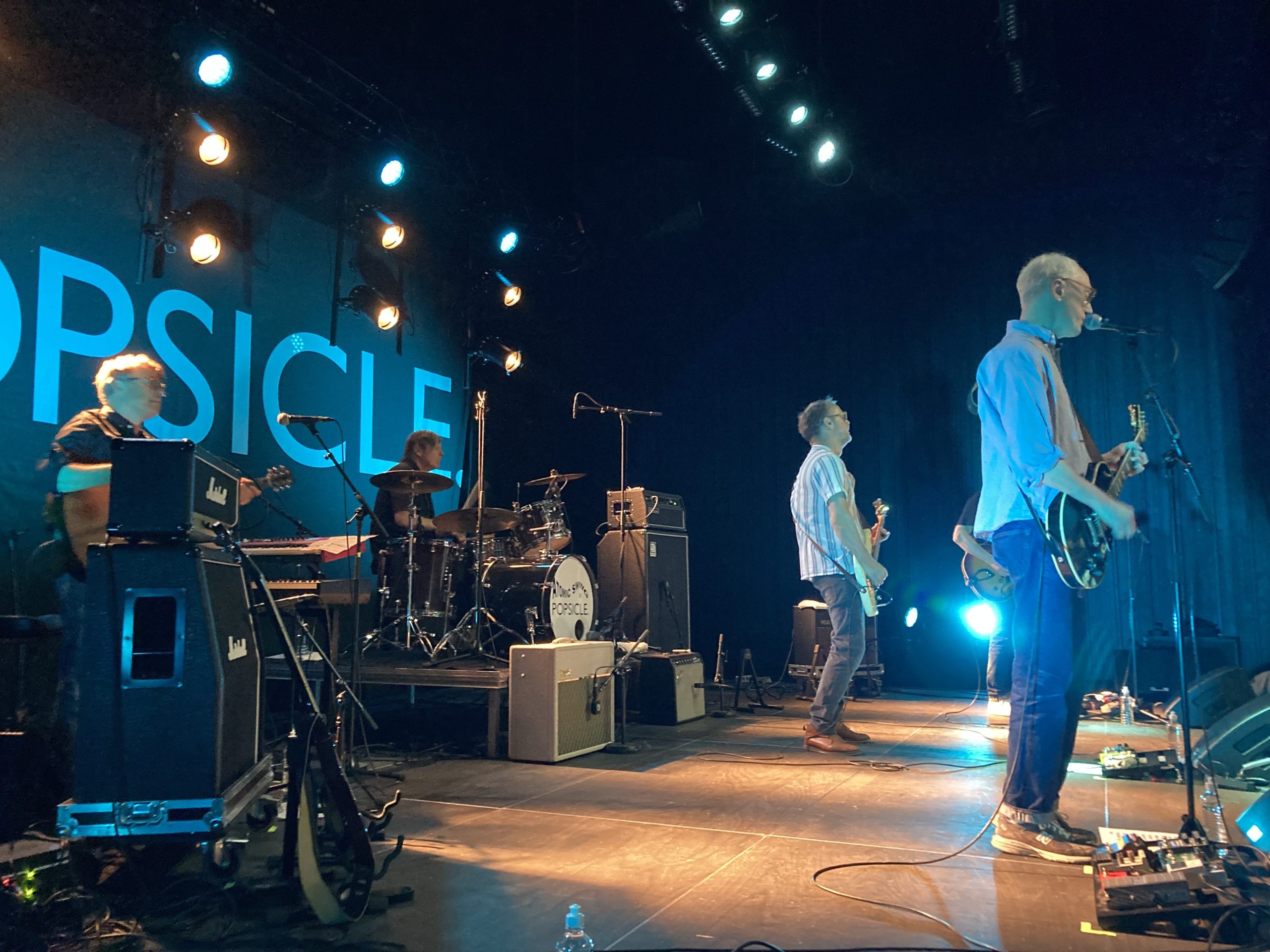 Popsicle på scen i blå scenbelysning. Fyra män i 50-årsåldern på trummor, akustisk gitarr, elbas och elgitarr. I förgrunden står Andreas Mattsson. Han har blå jeans och en ljusblå skjorta. Han spelar och sjunger i mikrofonen.