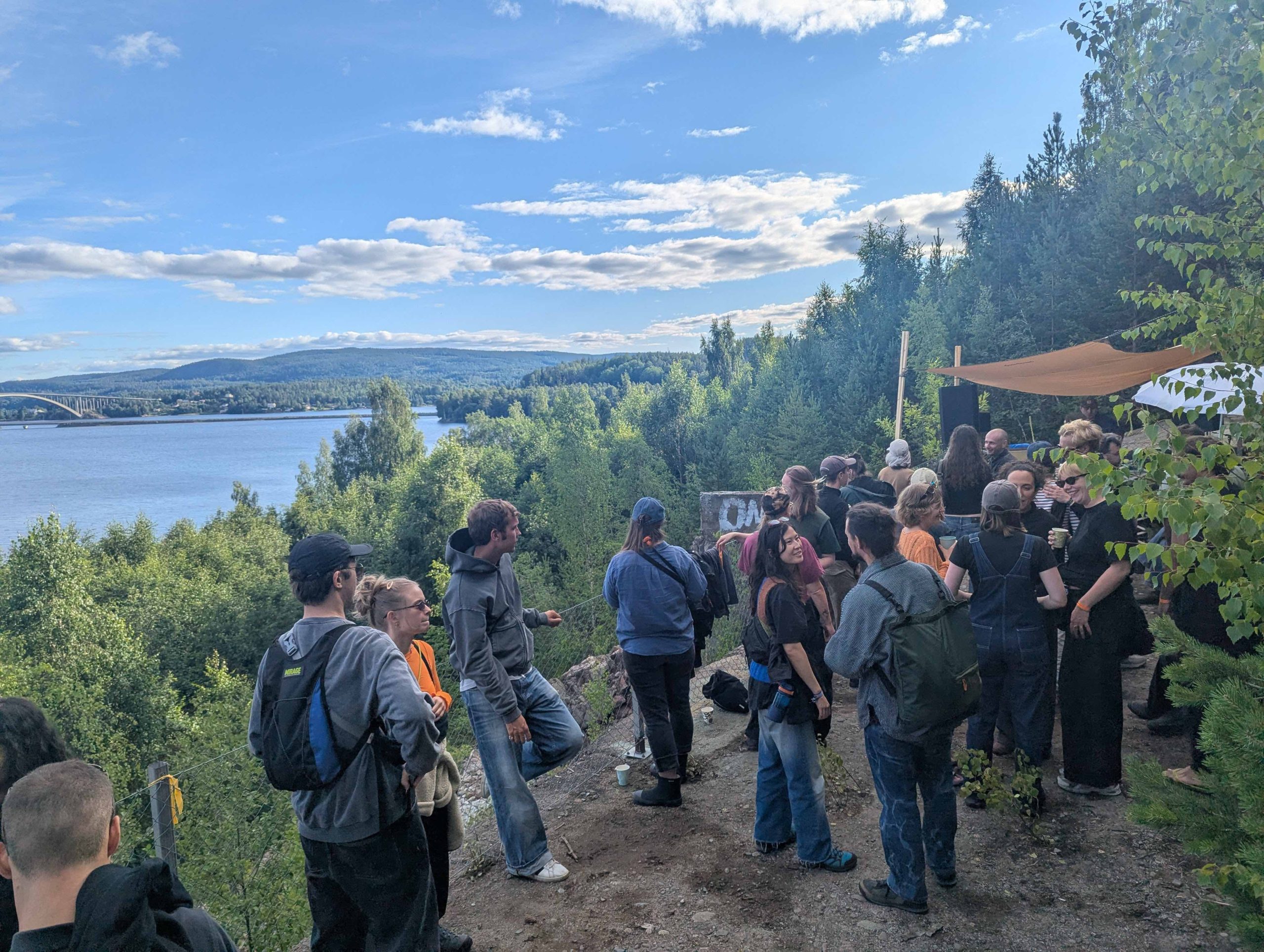 En folksamling som står på ett berg. Någon slags aktivitet verkar vara i görningen. I bakgrunden syns berg, en sjö och en bro.