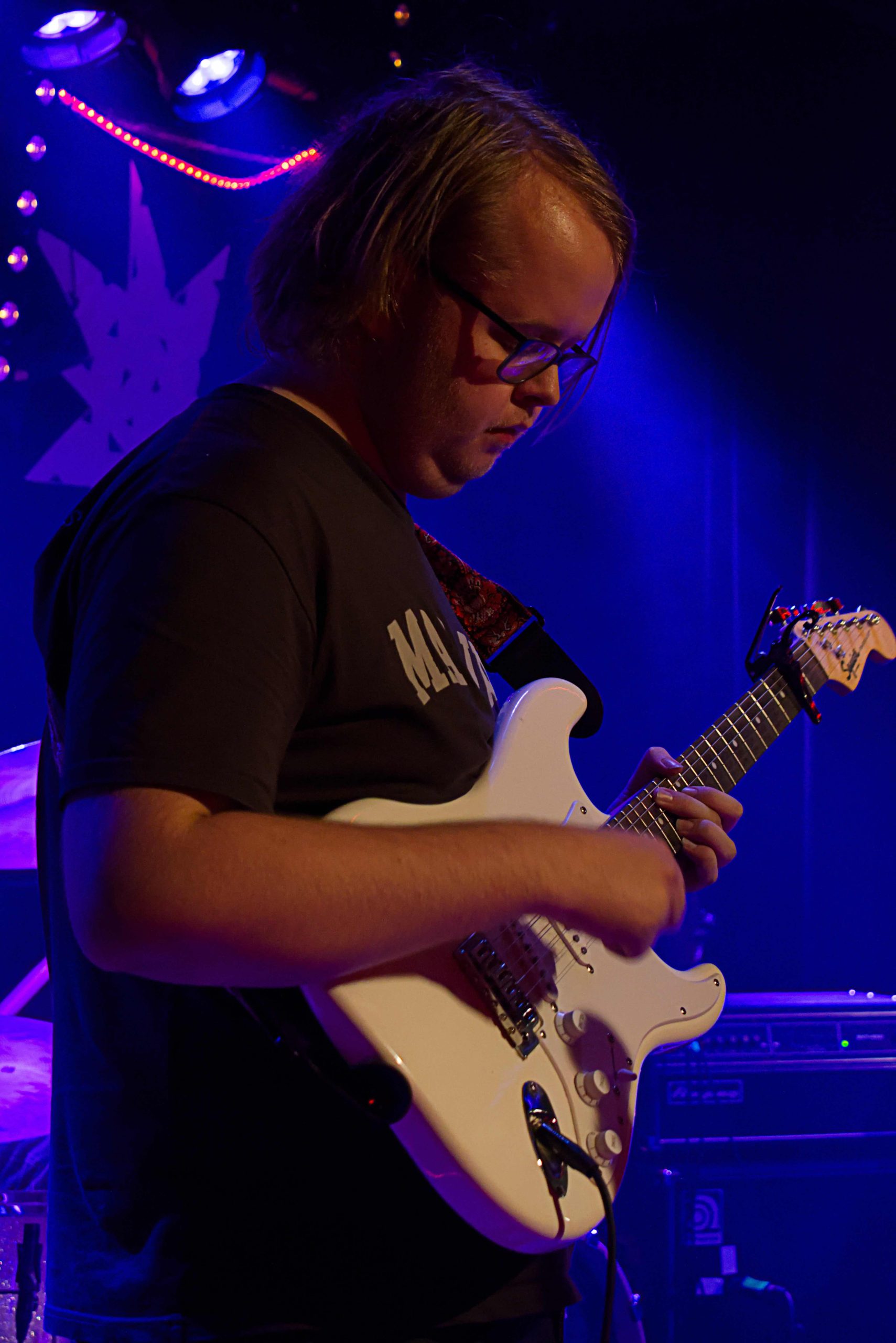 Carnal Flowers gitarrist spelar gitarr med huvudet sänkt och blicken på gitarren. Elgitarren är vit och gitarristen har mörk t-shirt, blont, halvlångt hår och glasögon.