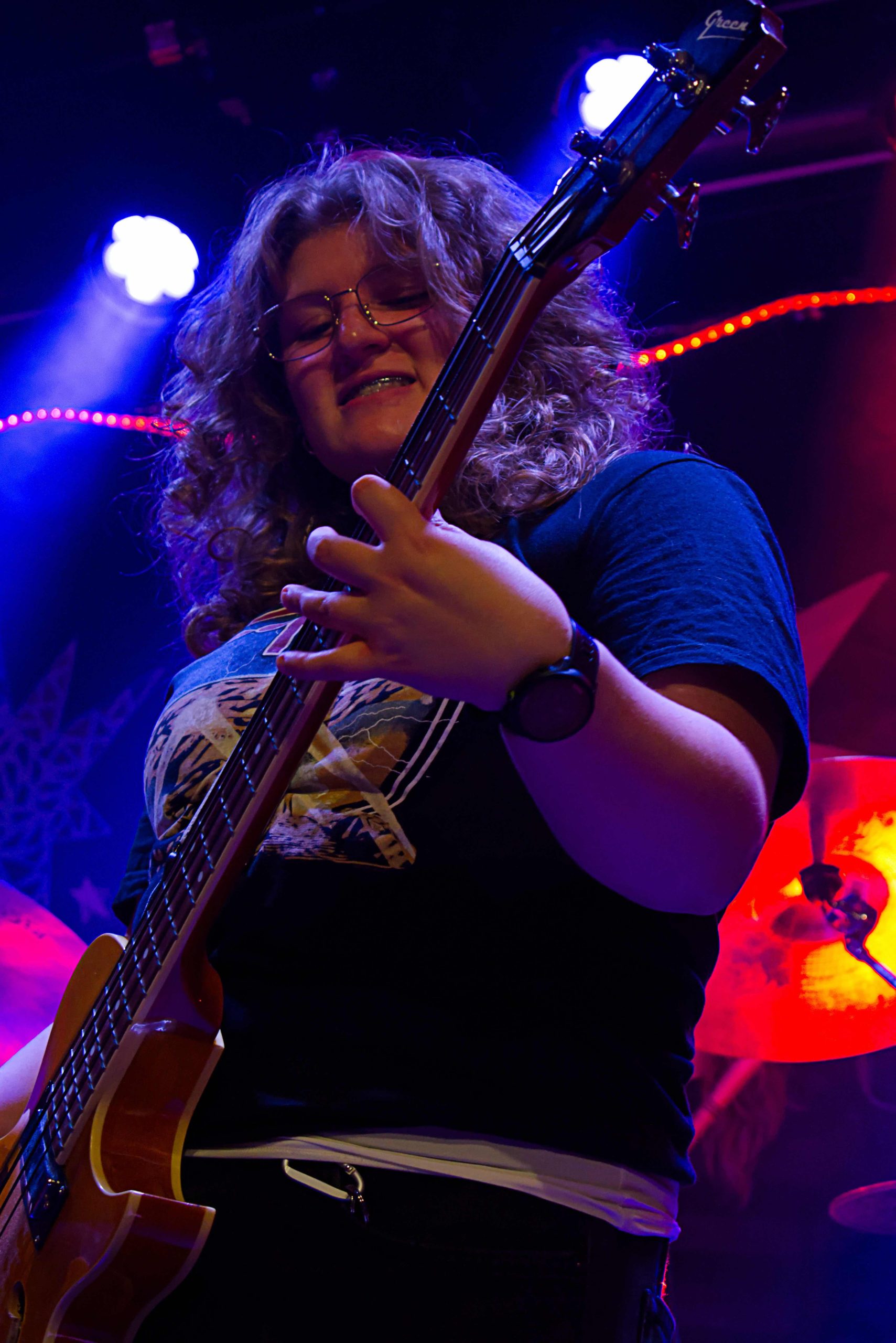 Gitarrist i Hållplats 11. Hon spelar med ett fokuserat ansiktsuttryck med blicken på gitarren. Hon har lockigt, ljust hår, glasögon och tandställning och en band-tshirt.