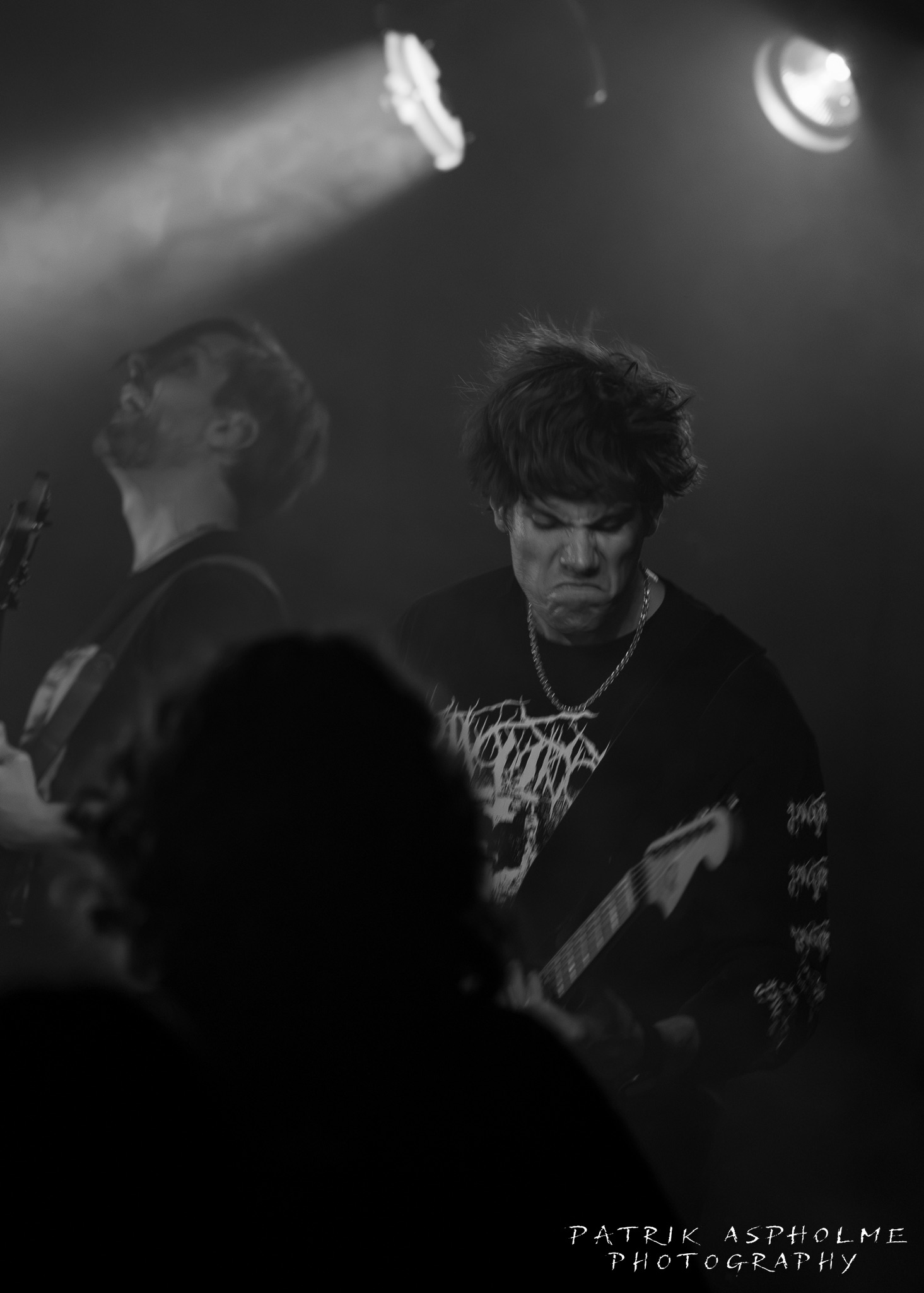 En man med förvridet ansiktsuttryck spelar gitarr. Han har svart bandtröja och mörkt, tjockt hår. Bakom honom syns den andra gitarristen.