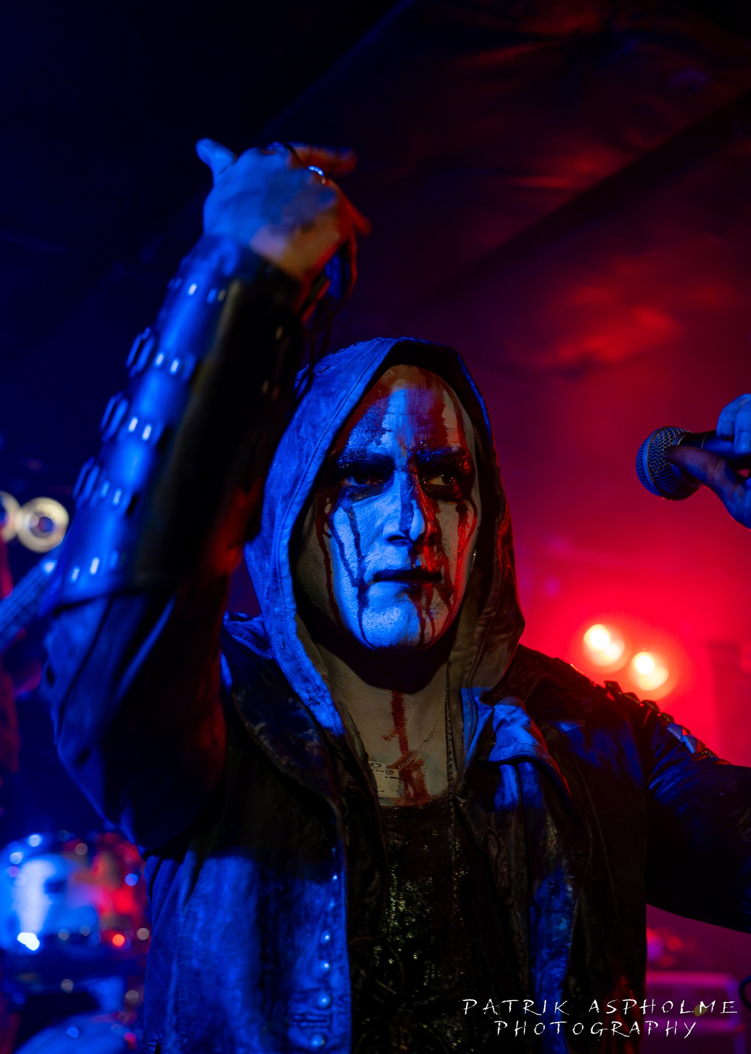 Nazghors frontman har luva, rinnande svart smink och fakeblod över hela ansiktet och ner mot halsen. Han höjer ena handen.