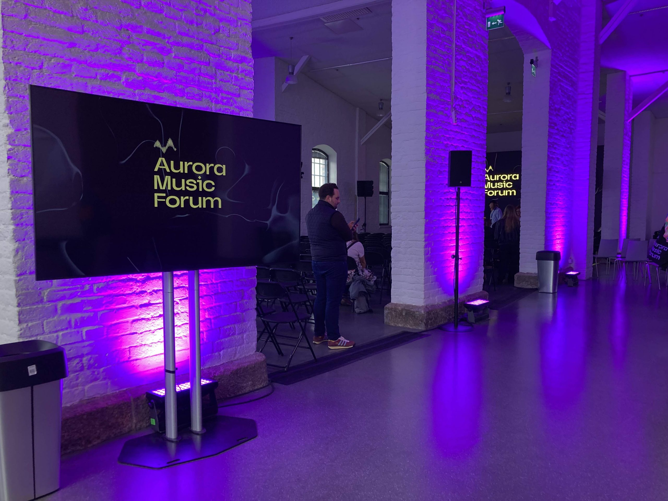 En av de stora salarna i tegelbyggnaden. En vitmålad sal med tegelväggar och lila belysning som färgar hela lokalen lila. Bilden visar en led-skärm med Aurora Music Forums logotyp.