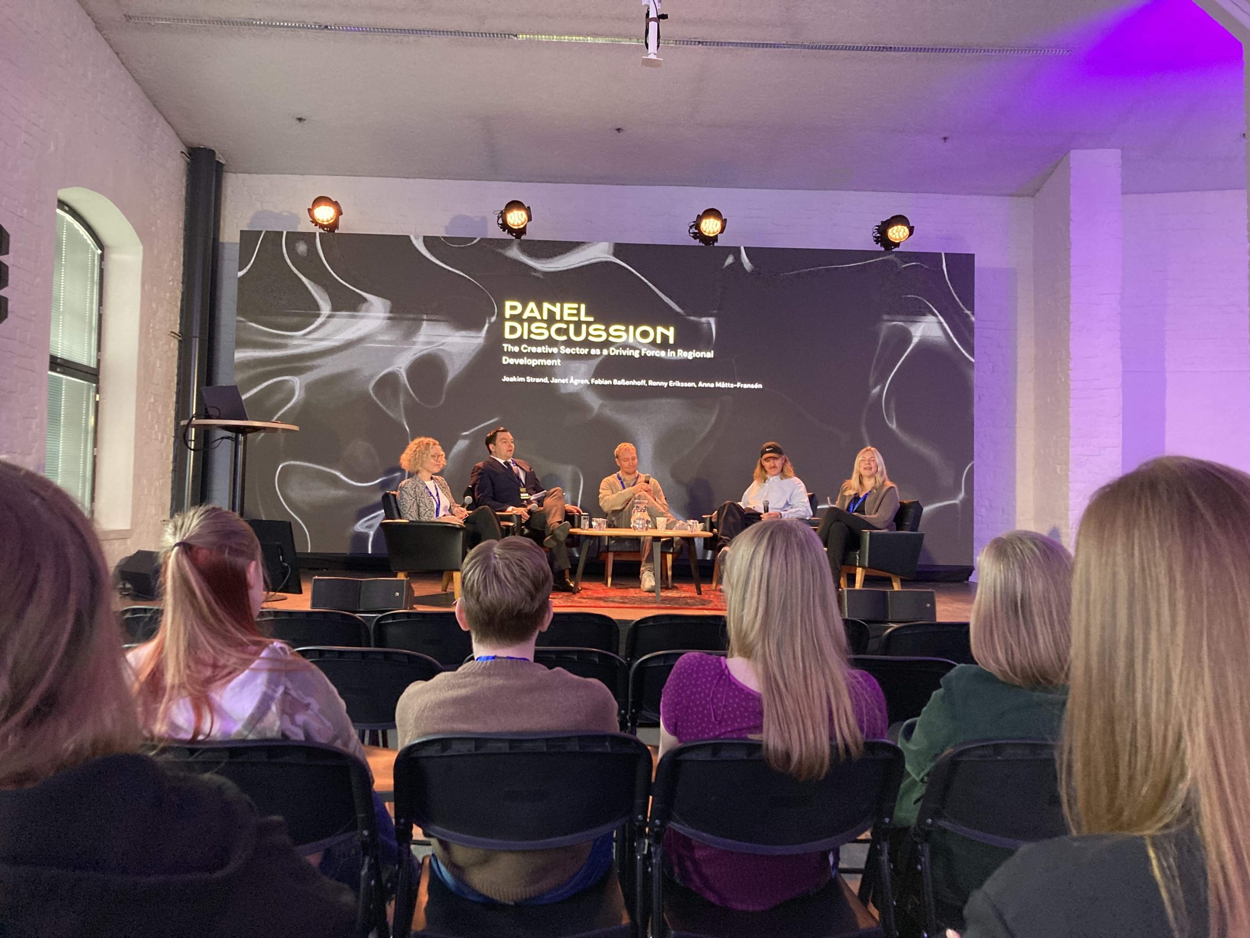En scen med fem personer som sitter i fåtöljer. Bakom dem hänger en stor ledskärm som visar vilken programpunkt det är: "Panel Discussion!