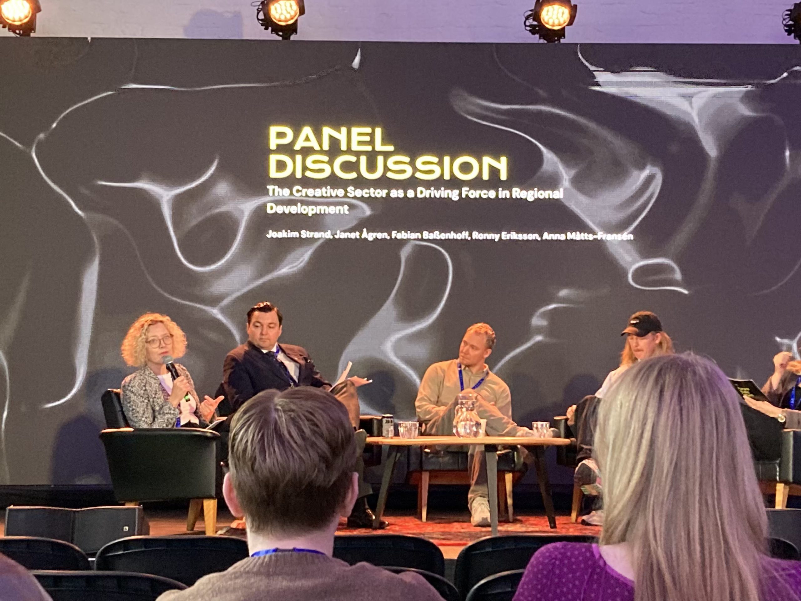 Panelsamtal med fem personer på scen. LED-skärmen bakom de säger "Panel Discussion". En kvinna i samtalet vänder sig mopt publiken medan hon talar i en mikrofon. Hon har rutig kofta/kavaj och blont, krusigt hår.