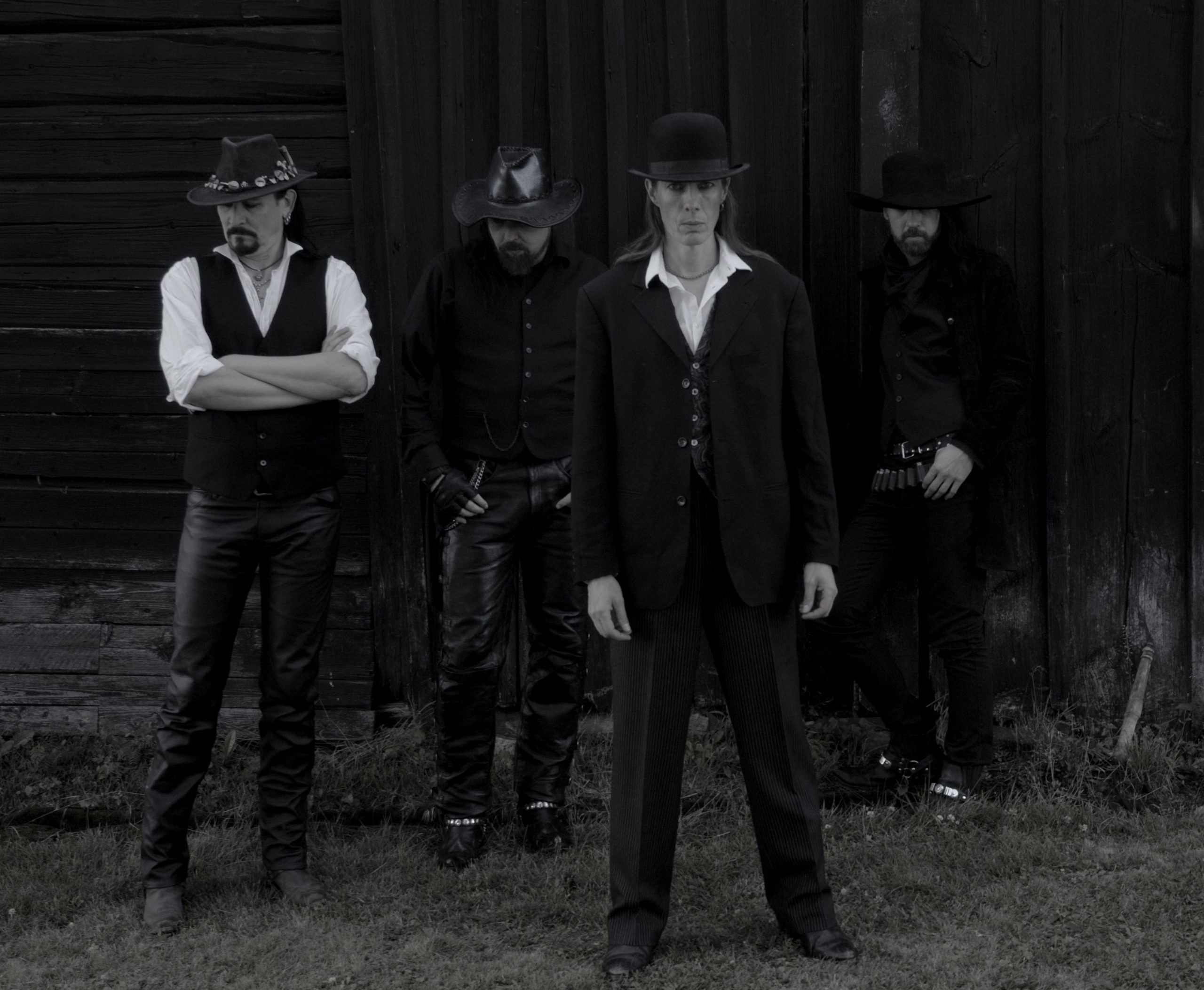 Fyra män poserar framför en laduvägg. Bilden är svartvit. Tre av männen bär cowboyhatt och mannen i förgrunden har ett plommonstp. De är propert klädda i väst, rock och kavaj. De ser mycket allvarliga ut.