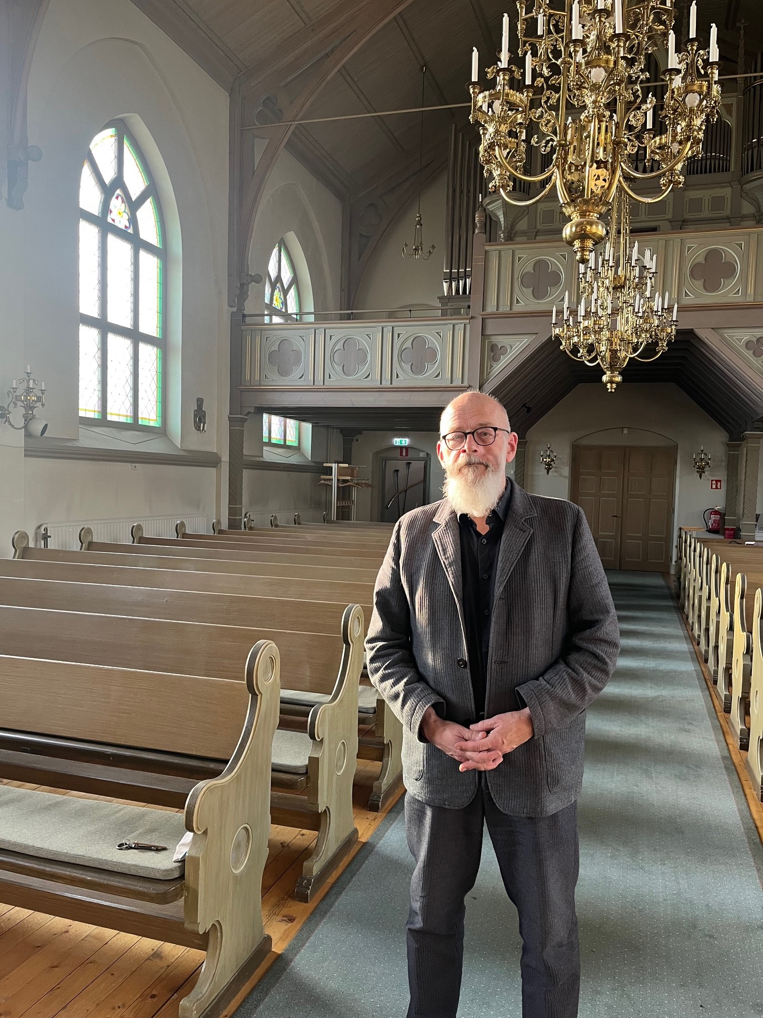 Hans-Ola i gången i Holms kyrka. Han ser in i kameran och har knäppta händer. Han har glasögon och grå kavaj. Han är skallig och har långt, vitt skägg. Kyrkans interiör är målat i blek gröngrått, den stora orgeln täcks delvis av två stora ljuskronor i guld.